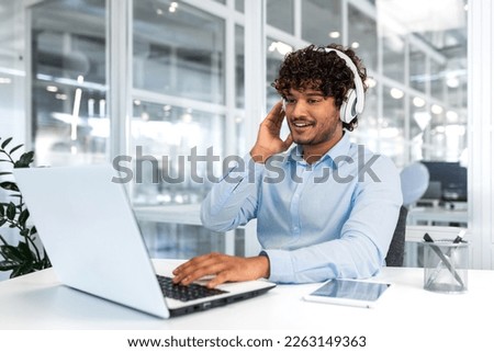 Young successful programmer inside modern office working with laptop, man in blue shirt smiling and happy, listening online music in headphones, audio books and podcasts, businessman satisfied work.