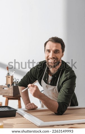 Cheerful restorer in apron holding paintbrush and looking at camera near wooden frame in workshop