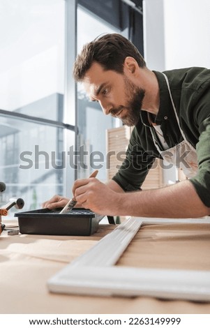 Bearded restorer holding paintbrush near paint and blurred wooden picture frame in workshop