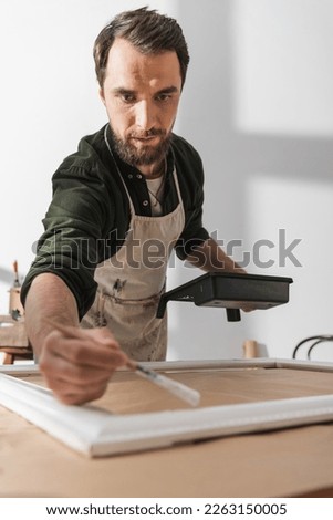 Focused restorer in apron holding paintbrush near blurred picture frame in workshop