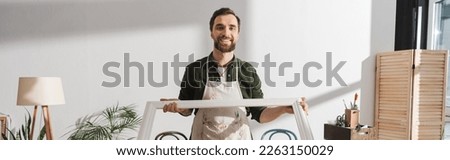 Smiling restorer holding wooden picture frame and looking at camera in workshop, banner
