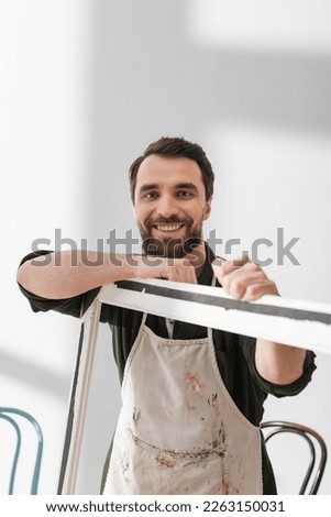 Cheerful restorer in apron looking at camera near wooden picture frame