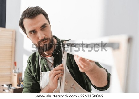 Bearded repairman in apron looking at blurred picture wooden frame in workshop