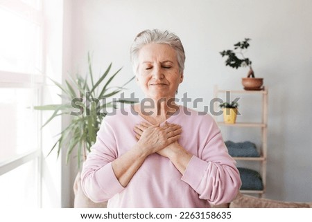 Portrait of happy adult elderly female volunteer holding folded hands on chest, close eyes. Kind smiling middle-aged mature woman feeling thankful, gratitude believe charity concept Royalty-Free Stock Photo #2263158635
