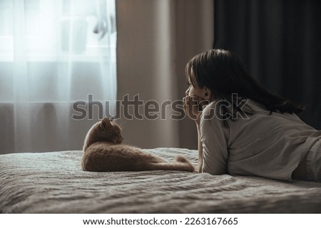 Sad young woman in pajamas with seasonal affective disorder lies alone on the bed near the window, next to the domestic cat. The concept of winter depression due to lack of sunlight Royalty-Free Stock Photo #2263167665