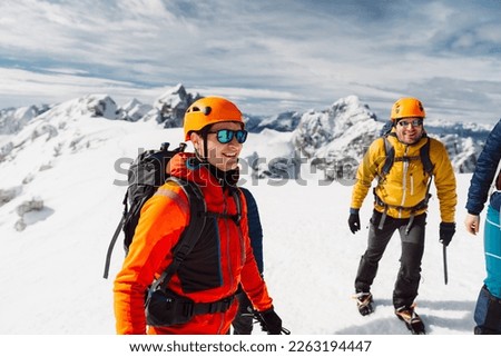 Mountaineer in orange jacket and protective helmet talking to the group while they enjoy the sun on top of the mountain