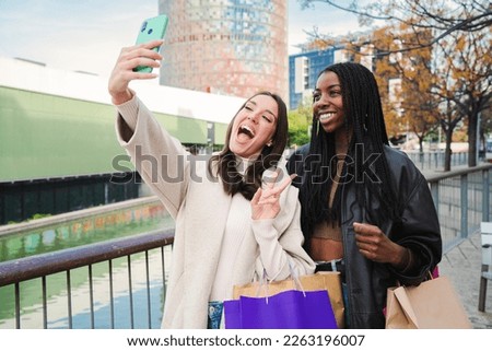 Two multiethnic young women smiling and having fun taking a selfie portrait with a smart phone after shopping and sharing in the social media. A couple of carefree girls doing a photo with a cellphone
