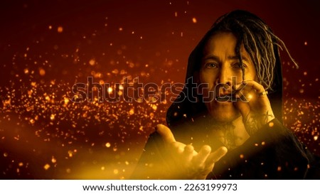 A male shaman in ethnic clothing with a hood, inviting you into his mysterious world. Dark red background with sparks and copy space. Halloween, Fantasy. Royalty-Free Stock Photo #2263199973
