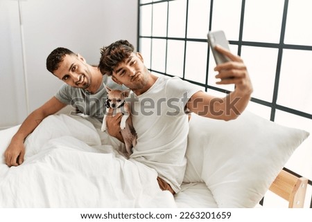 Two hispanic men couple make selfie by the smartphone sitting on bed with chihuahua at bedroom