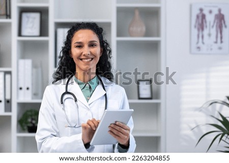 Portrait of successful smiling hispanic female doctor inside medical office, woman with tablet computer close up smiling and looking at camera, working in medical gown while standing. Royalty-Free Stock Photo #2263204855