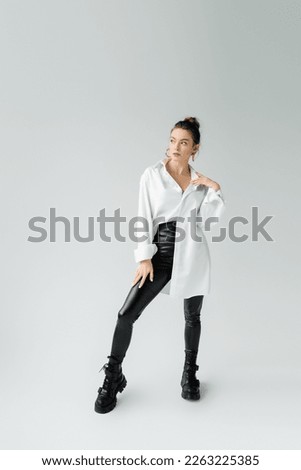 full length of trendy woman in oversize shirt and tight pants with rough boots looking away on grey background Royalty-Free Stock Photo #2263225385