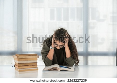 Young girl future law or economics student feeling nervous and anxious because she has a lot to study for her college entrance exam sitting with a pile of books on the table and has a headache
