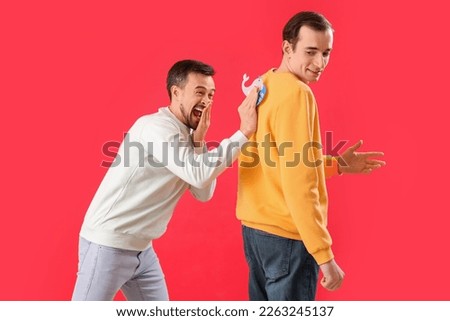 Young man sticking paper on his friend's back against red background. April Fools' Day celebration Royalty-Free Stock Photo #2263245137