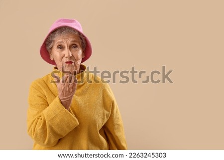 Senior woman in bucket hat blowing kiss on beige background