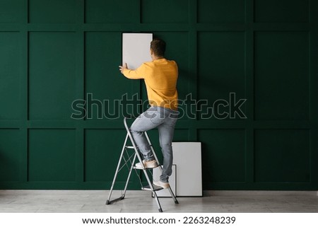 Young man with ladder hanging frame on green wall, back view