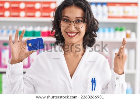 Young hispanic woman working at pharmacy drugstore holding credit card doing ok sign with fingers, smiling friendly gesturing excellent symbol 