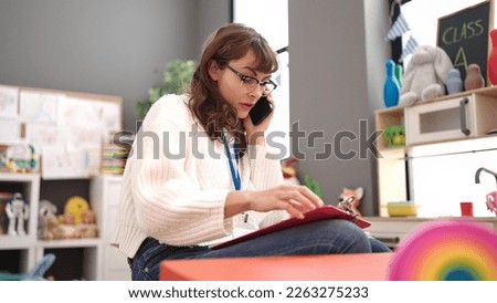 Young caucasian woman working as teacher writing on clipboard speaking on the telephone at kindergarten