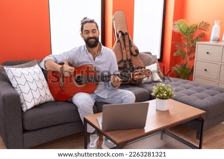 Hispanic man with beard playing classic guitar at home smiling happy and positive, thumb up doing excellent and approval sign 