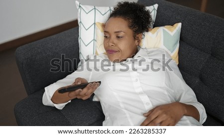 African american woman watching tv lying on sofa with boring expression at home