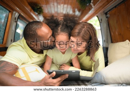The back of a white classic van, little kids play and learn about the world on their tablet with parents taking care of helping.