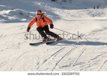 A young female skier in an orange jacket turns carving arches on the slope. A beautiful sunny day in the mountains, on the ski slope, a skier in an orange jacket is going down a black slope. Italy