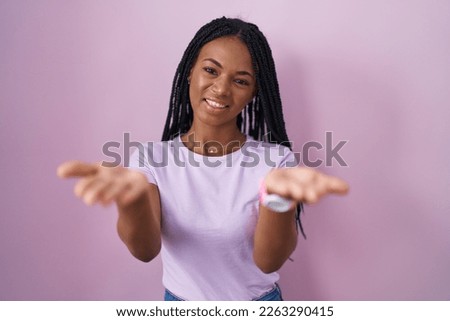African american woman with braids standing over pink background smiling cheerful offering hands giving assistance and acceptance. 