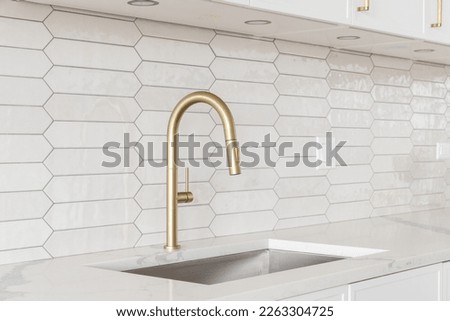 A beautiful kitchen faucet detail with white cabinets, a gold faucet, white marble countertops, and a brown picket ceramic tile backsplash. Royalty-Free Stock Photo #2263304725