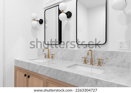 A bathroom with a white oak cabinet, marble countertop, gold faucets, black and white tiled floor, and sconces around black mirrors. Royalty-Free Stock Photo #2263304727