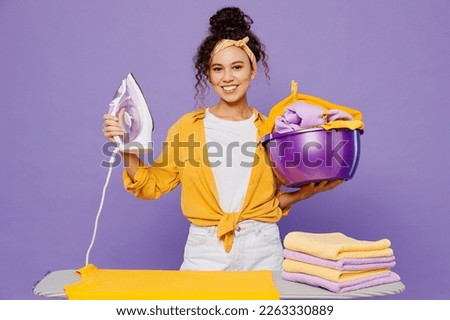 Young fun housekeeper woman wear yellow shirt white t-shirt tidy up ironing clean clothes on board hold basin with garment isolated on plain pastel light purple background studio. Housework concept