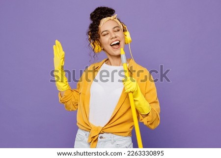 Young housekeeper woman wear yellow shirt rubber gloves headphones listen music dance tidy up hold broom like microphone sing song isolated on plain pastel purple background studio. Housework concept