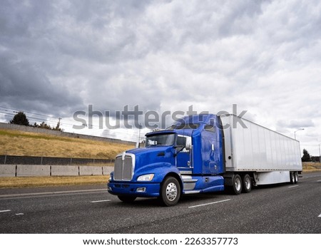 Classic dark blue big rig American popular bonnet semi truck with dry van semi trailer going on wide highway with stormy sky caring commercial cargo for delivery to business warehouse  Royalty-Free Stock Photo #2263357773