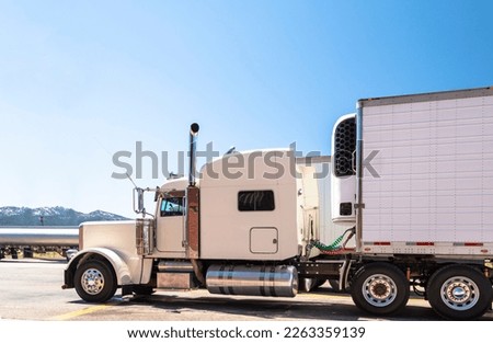 White big rig beautiful classic American powerful semi truck with chrome exhaust pipes with attached refrigerated semi trailer with refrigerator unit on it standing on truck stop parking lot in Utah Royalty-Free Stock Photo #2263359139