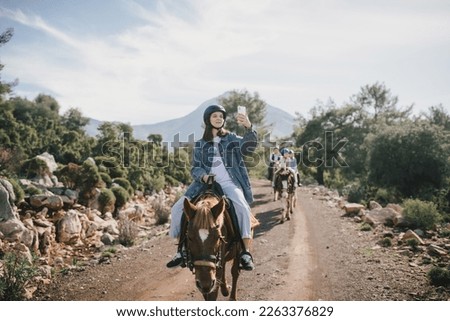 A young woman takes photos of a group of people on her smartphone  riding a horse through picturesque landscapes.