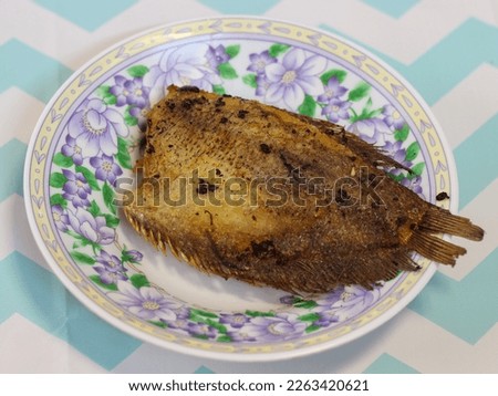 Deep-fried gourami lies on a white plastic plate with a floral pattern on a colorful tablecloth as a background