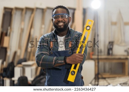 Portrait of African American male carpenter holding precision level making new furniture at wood workshop. Portrait of joiner male working with precision level in wood factory Royalty-Free Stock Photo #2263436427