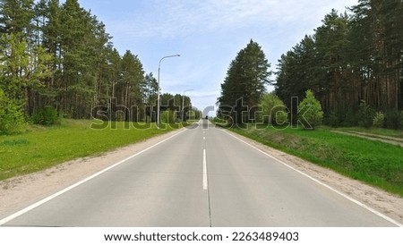 A concrete road with markings runs through a wooded area. Along the sides of the road are grassy lawns. There are lamp posts along the road. Blue sky with clouds Royalty-Free Stock Photo #2263489403