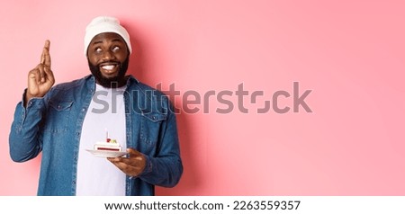 Hopeful Black guy celebrating birthday, making wish with fingers crossed, holding bday cake with candle, standing against pink background.