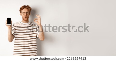 Technology and e-commerce concept. Young man with red hair showing okay sign and blank smartphone screen, praising awesome app or video game, white background.