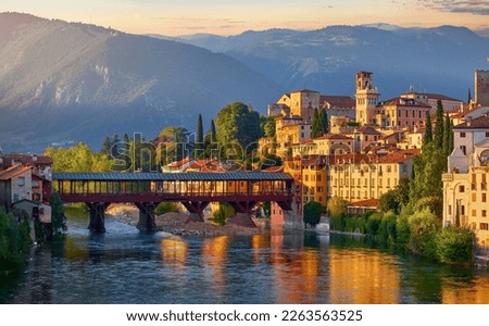 Bassano del Grappa, Veneto, Italy. Bridge Ponte degli Alpini at river Brenta. Panoramic view old town with vintage building tower, wooden Alpine mountains scenic sunset landscape Royalty-Free Stock Photo #2263563525