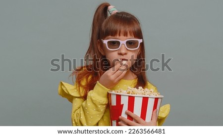 Excited young toddler school girl in 3D glasses eating popcorn, watching interesting tv serial, sport game, film, online social media movie content. Teen female child kid on studio gray background
