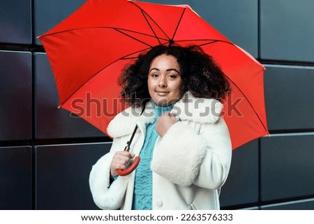 Fashion portrait of young chubby woman holding a red umbrella against black wall in London. She is wearing white coat and blue sweater. Royalty-Free Stock Photo #2263576333
