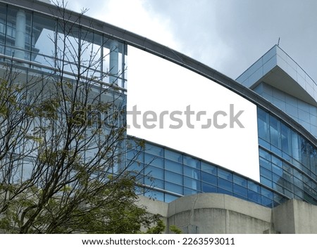 Blank advertising horizontal banner poster mockup on modern building with glass facade exterior, trees on the left. Super large billboard, out-of-home OOH media display space.