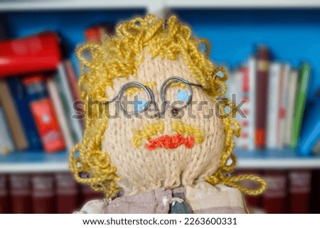 Knitted and crocheted male doll with mustache , glasses , blue eyes and blond curly hair . Close-up portrait . Man in front of blurred bookshelf . Doll in the shape of a nerdy engineer . Up cycled DIY