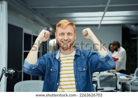 Young red-haired programmer or businessman in modern office looking at camera and smiling. Freelancer celebrating a business success with arms raised in meeting room. Colleagues work in background.