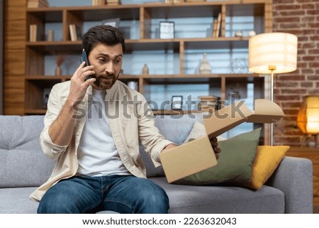Angry cheated man at home with phone and boxed parcel received, arguing on phone talking to online store customer support service, product delivery failed. Royalty-Free Stock Photo #2263632043