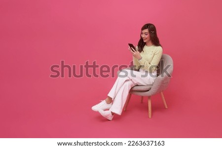 Happy young Asian woman using mobile phone. While her sitting on chair isolated on pink studio copy space background.