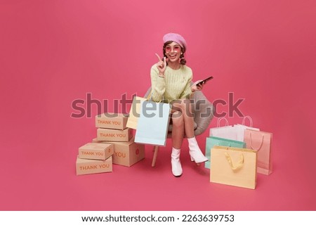Happy Asian teen woman sitting on sofa holding shopping bags and smartphone isolated on pink background, Shopper or shopaholic concept. Royalty-Free Stock Photo #2263639753