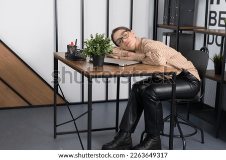 This photo captures a peaceful moment in a busy office. A young woman wearing glasses and a beige shirt takes a nap at her desk with a notebook, plant, and stationery items.