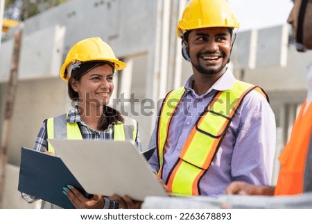 Team of construction engineers wearing vest and helmet safety discussing project at construction site.Group Indian foreman with laptop, paperwork working at factory making precast concrete wall.