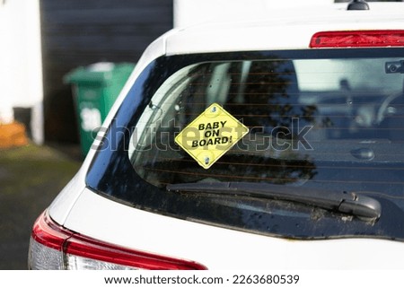 "BABY ON BOARD!" small yellow warning sign street sign suction cupped to the inside of a back windshield of a white family car in front of a driveway with a garbage or recycling bin in an English town